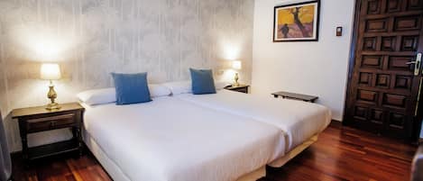 A double bed with white linens, two nightstands with lamps, a wooden floor, a wall-mounted air conditioner, and a door with a geometric pattern.