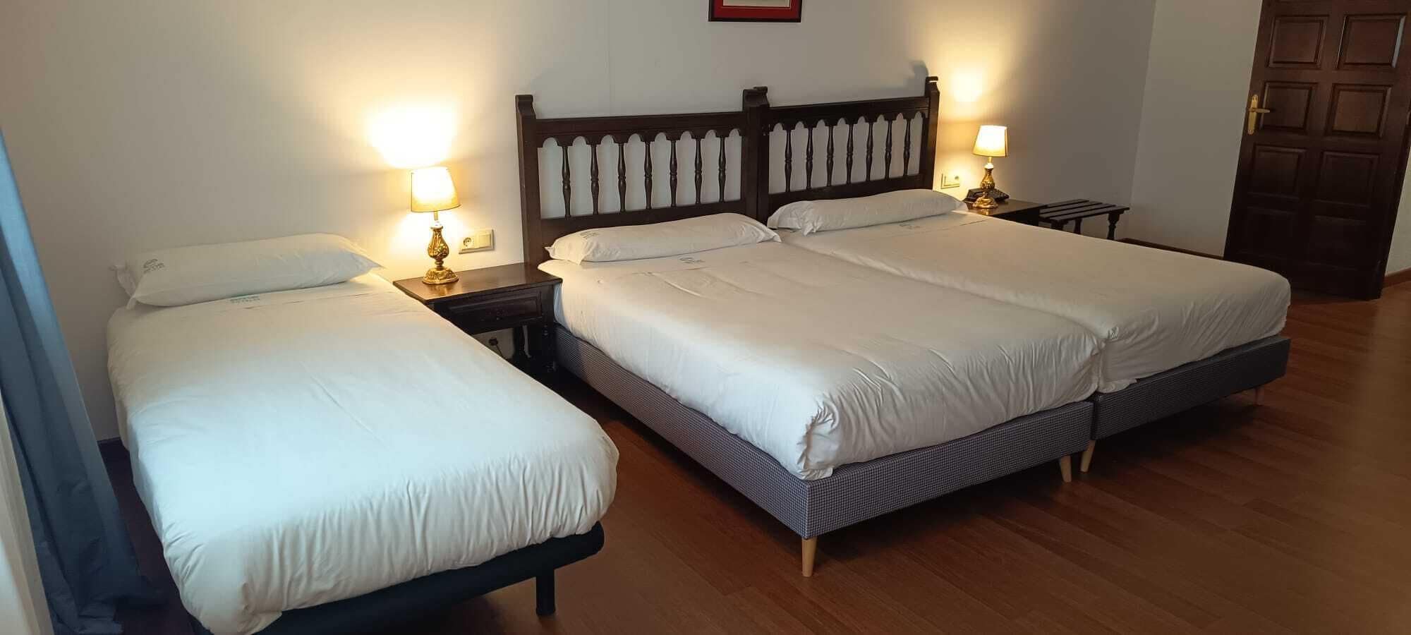 A hotel room with two single beds, wooden headboards, bedside lamps, and a wooden floor.