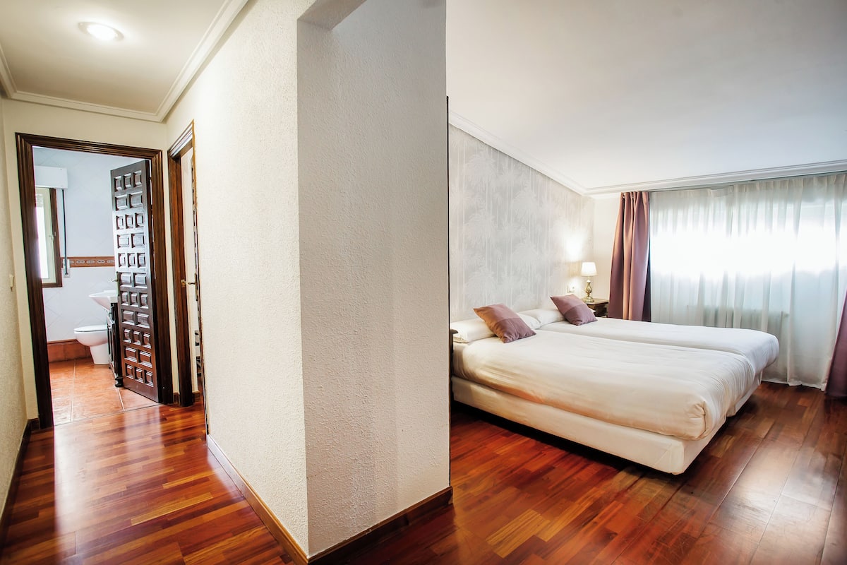 Habitació d'hotel amb un sòl de fusta, un llit gran amb almoades, una tauleta de nit i una finestra amb cortines.