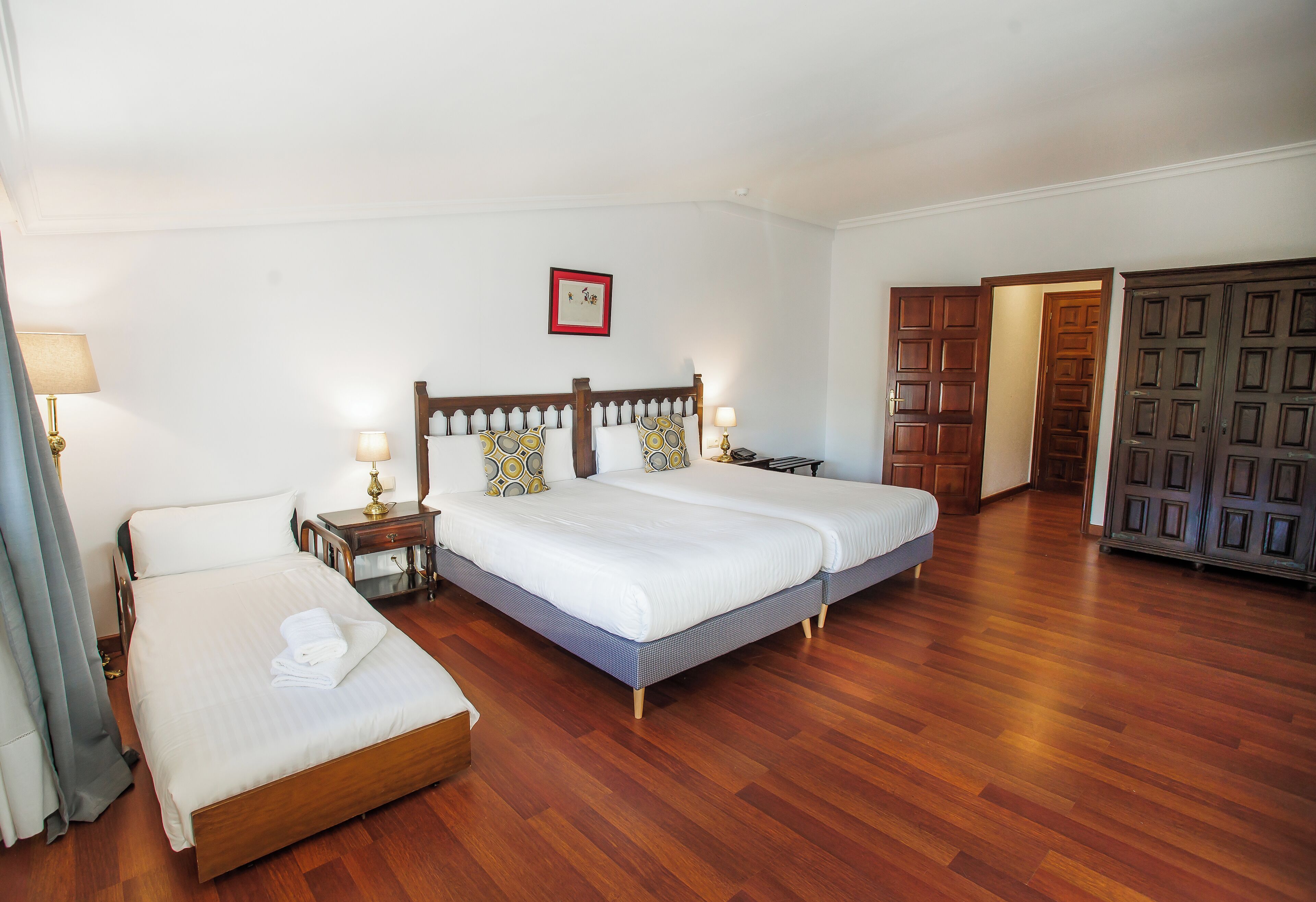A hotel room with two beds, a wooden floor, a bedside table with a lamp, and a wardrobe.