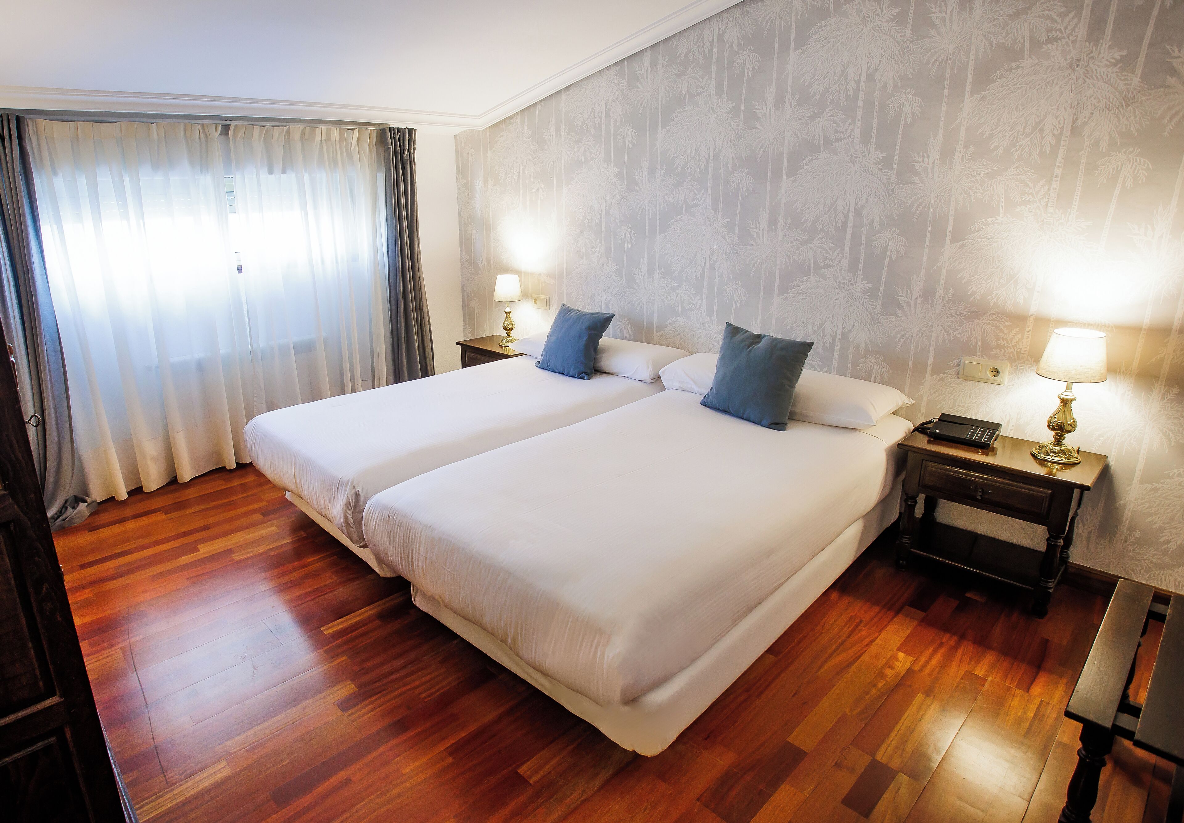 A double bed with two pillows, a bedside table with a lamp, and a wooden floor.