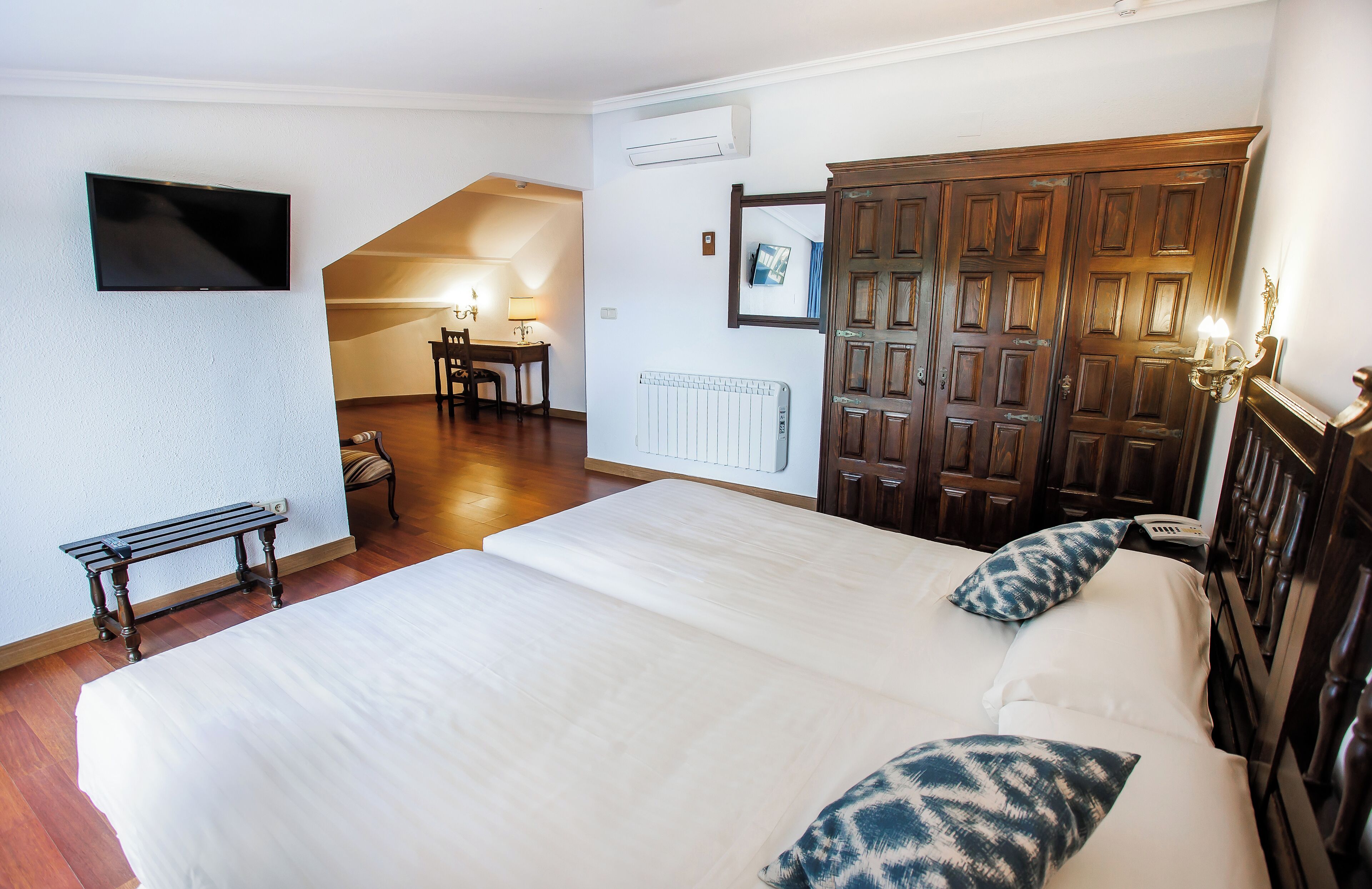 Habitación de hotel con una cama grande, un escritorio con silla, un radiador, un televisor montado en la pared y un armario de madera.