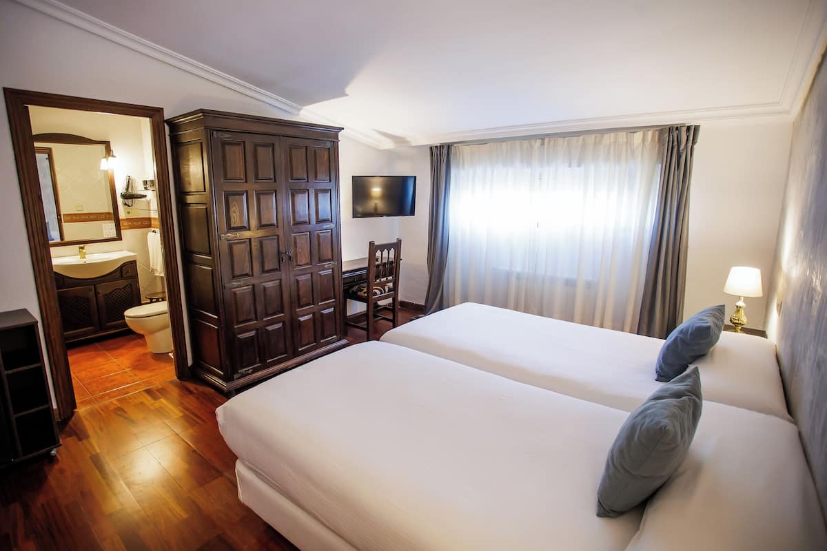 Habitació d'hotel amb un armari de fusta, televisió, llit amb almoades, bany amb rentapla i vàter, i finestra amb cortines.