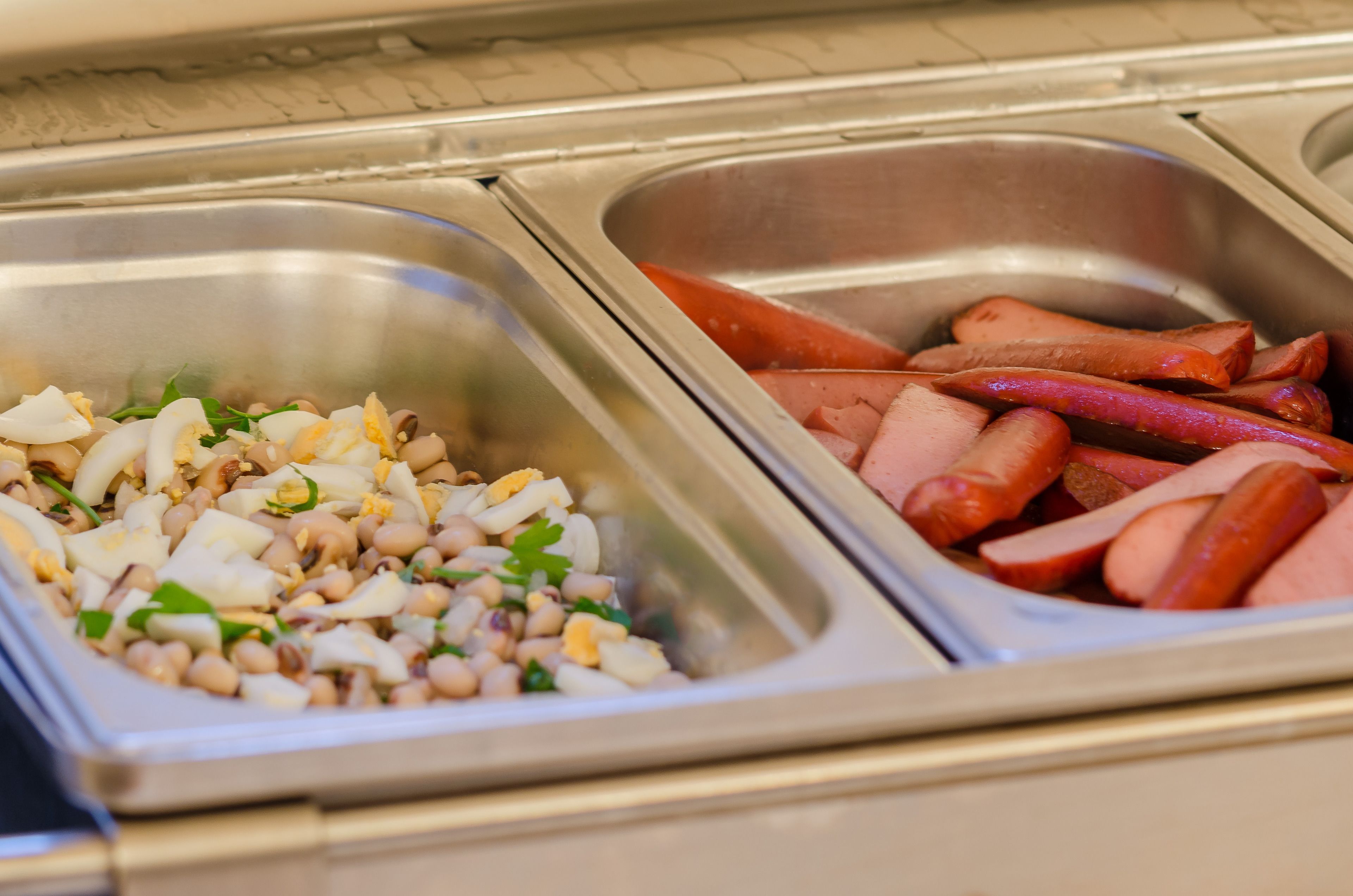 Een buffet met twee roestvrijstalen opschepkommen, waarvan er één belegd is met plakjes worst en de andere met een salade met bonen, eieren en bladgroenten.