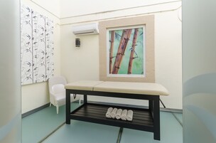 Ένα μικρό, καθαρό δωμάτιο με μια λευκή καρέκλα, ένα παγκάκι με ένα ράφι από κάτω και ένα κλιματιστικό τοποθετημένο στον τοίχο.
