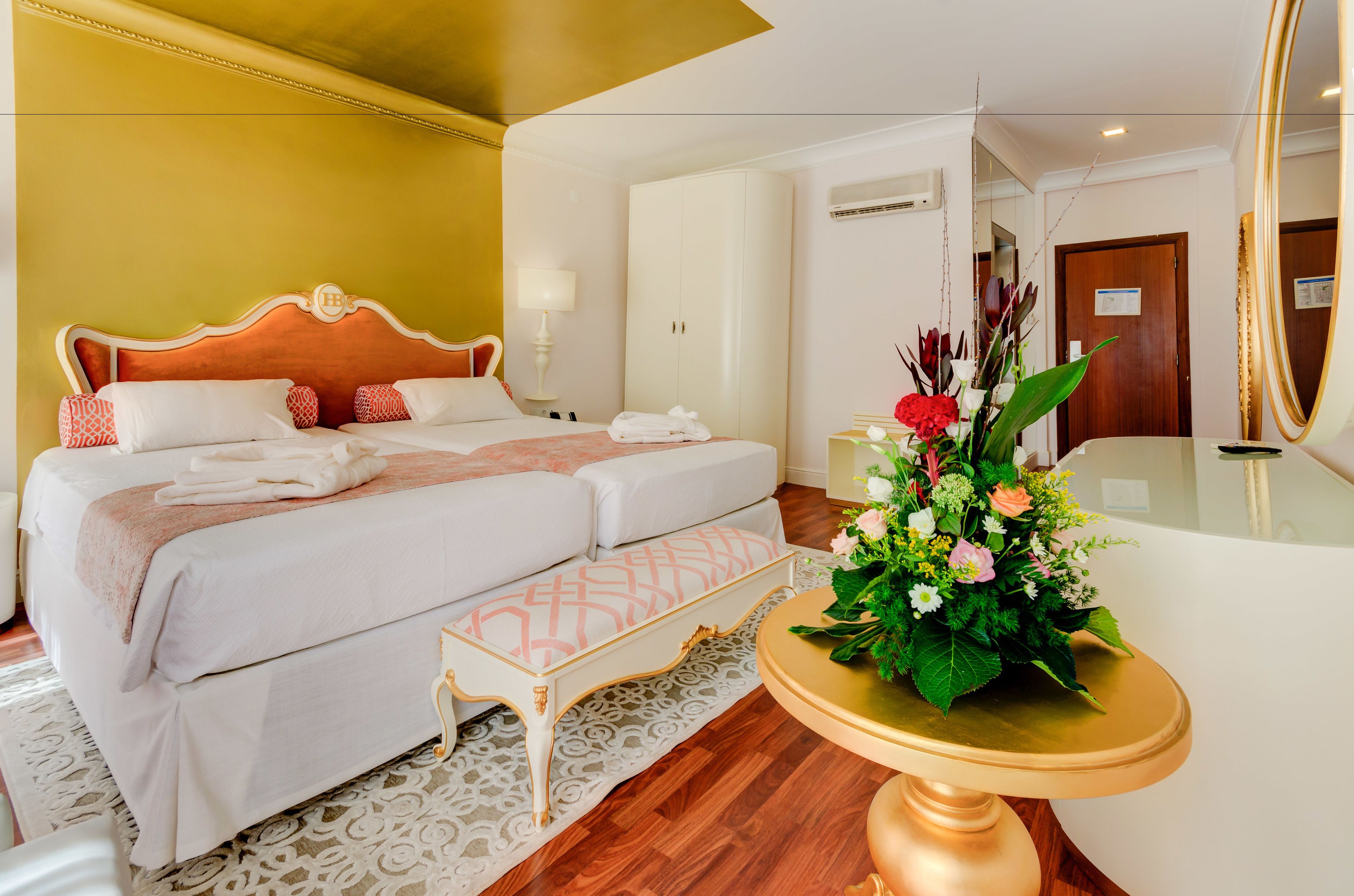 Hotelkamer met een tweepersoonsbed, nachtkastje met bloevas, een klein bankje en een grote spiegel.