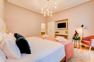 A hotel room with a large bed, a TV mounted on the wall, a pink armchair, and a small table with a flower arrangement.
