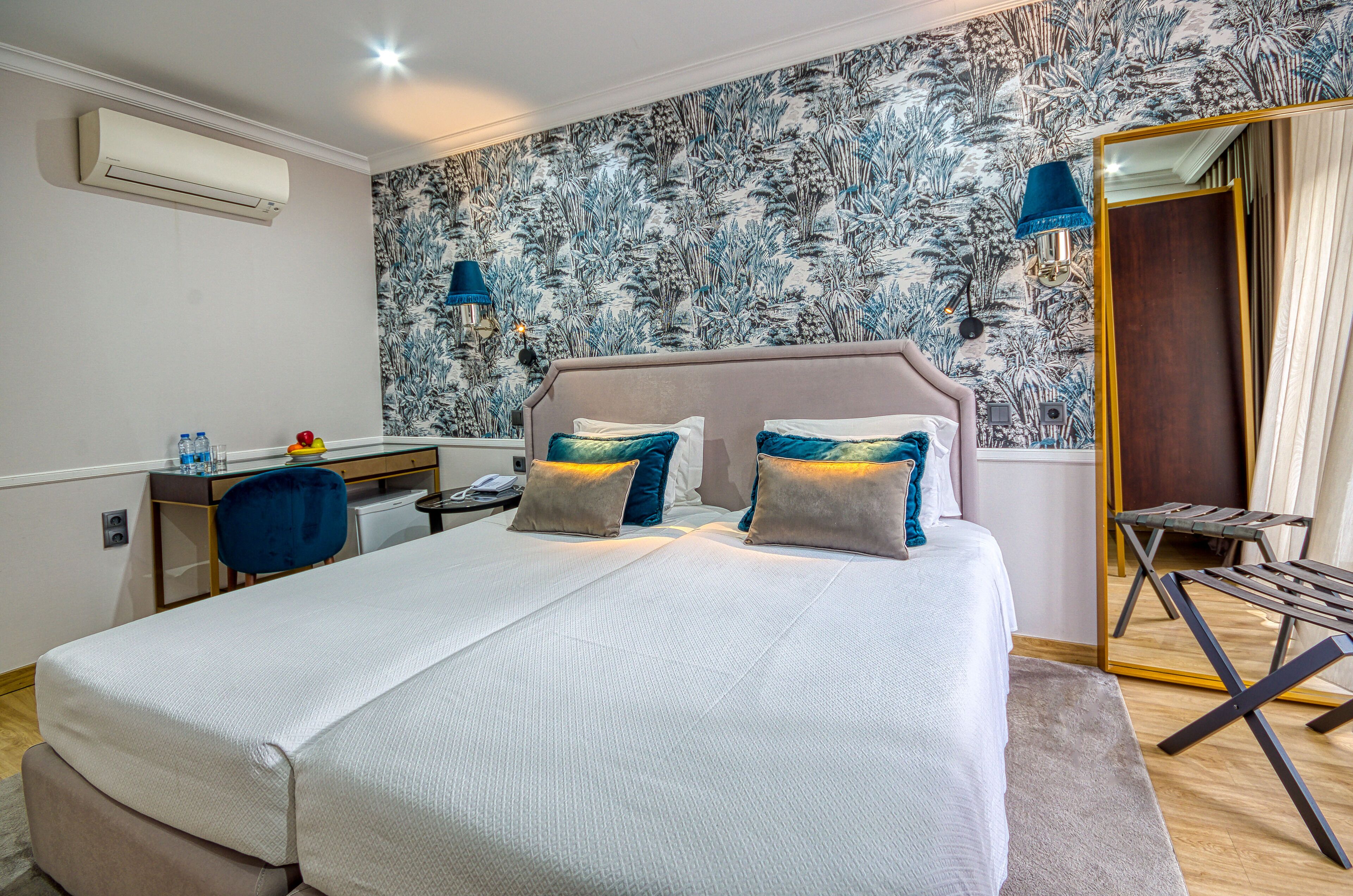Une chambre d’hôtel avec un grand lit, un bureau et une chaise. On y trouve un miroir, une lampe et une fenêtre.