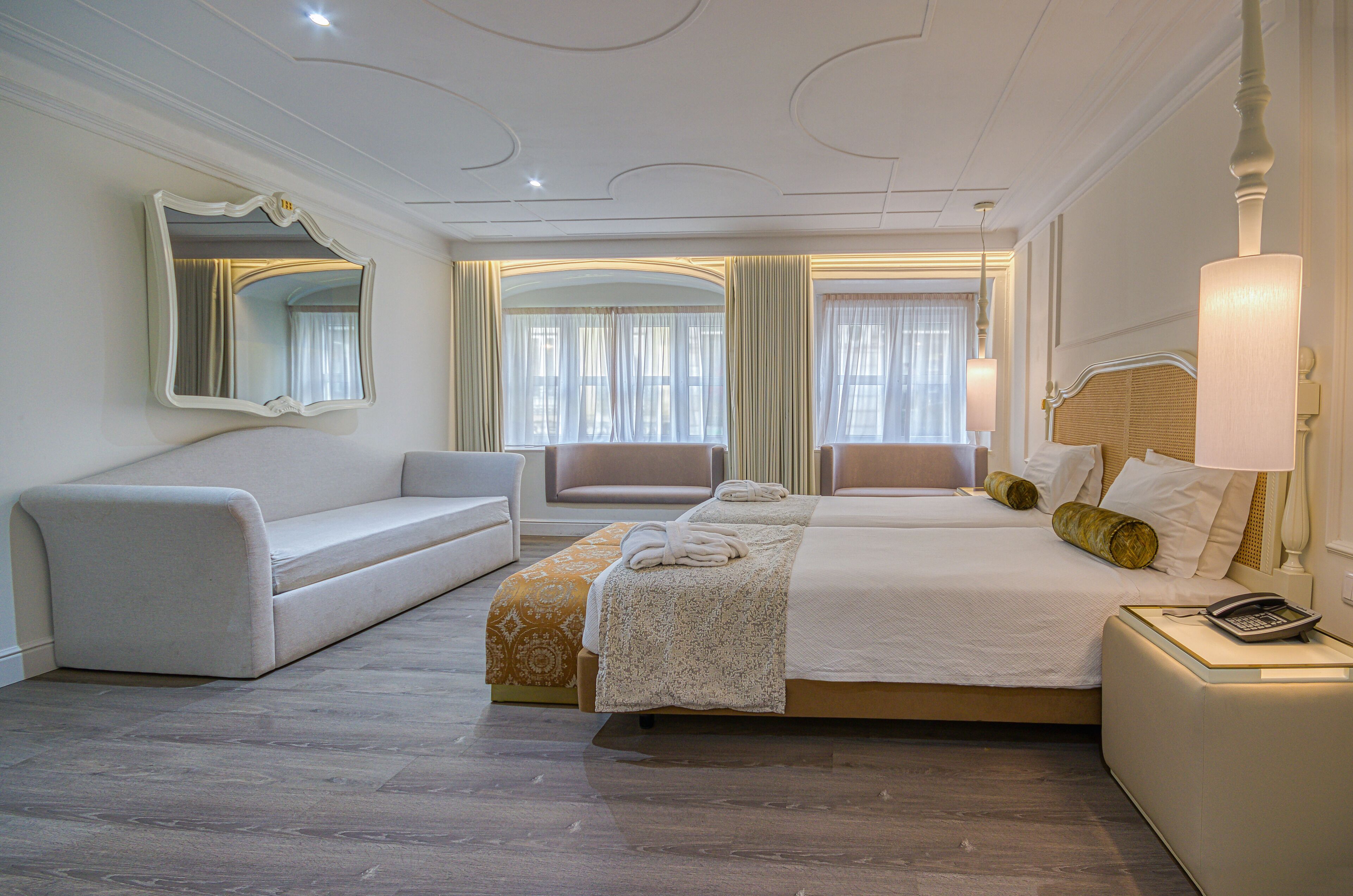 A hotel room with a large bed, a sofa, a chair, and a bedside table with a lamp.