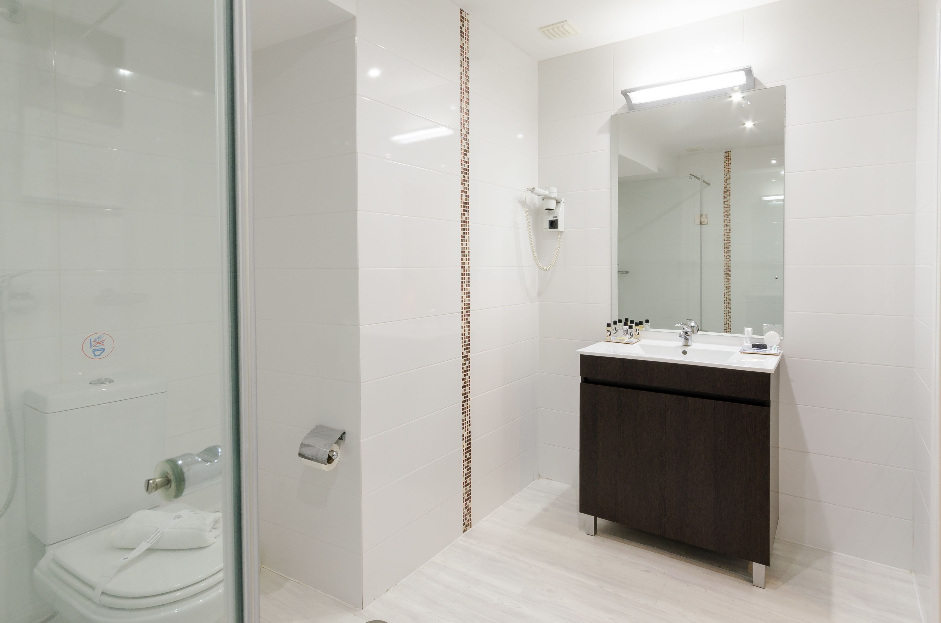 A modern bathroom with a glass shower enclosure, a toilet, a sink with a dark cabinet, and a mirror with a light above it.