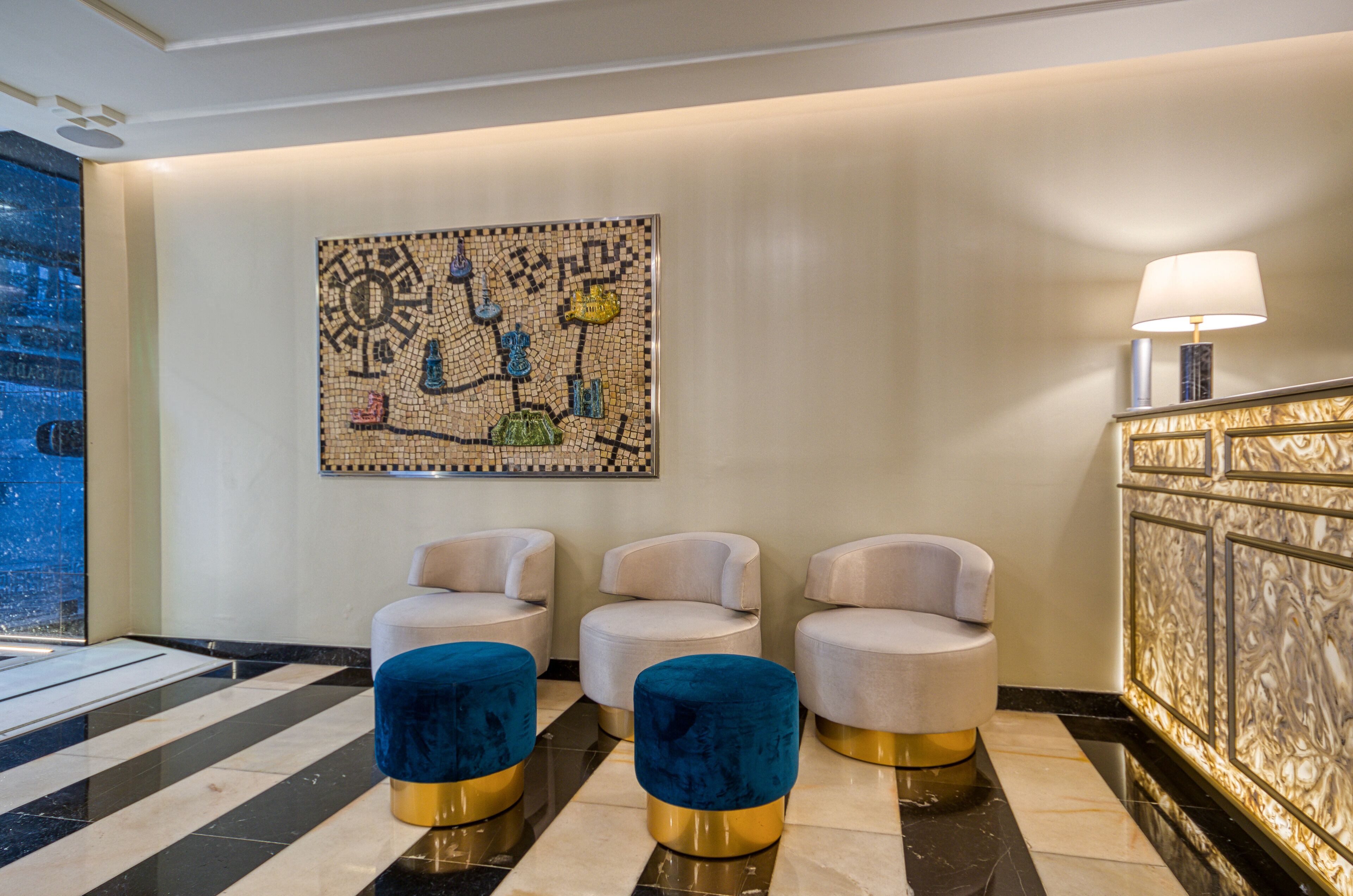 Un hall con tres sillones blancos, un reposapiés azul, una obra de arte de mosaico y un suelo de mármol.