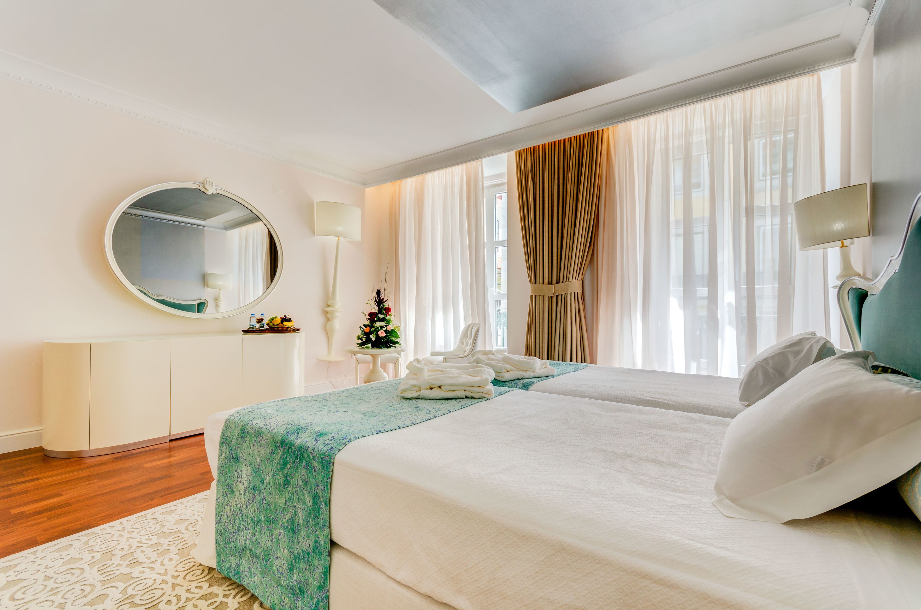 Camera d'albergo con due letti, uno specchio rotondo, un comodino con una lampada, una piccola pianta e vista all'esterno attraverso tende leggere.