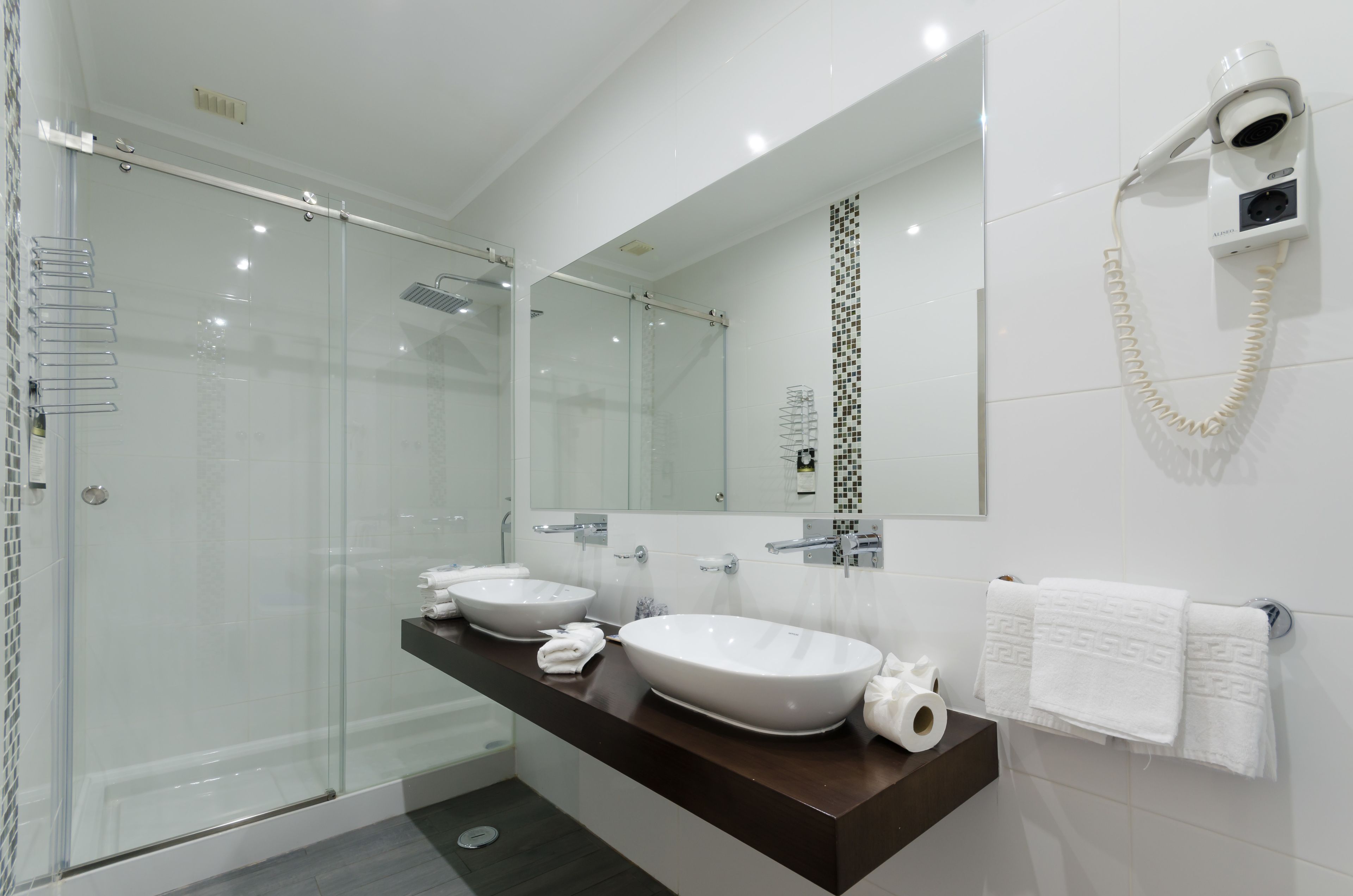 A modern bathroom with a glass shower enclosure, two sinks, a large mirror, and a hairdryer mounted on the wall.