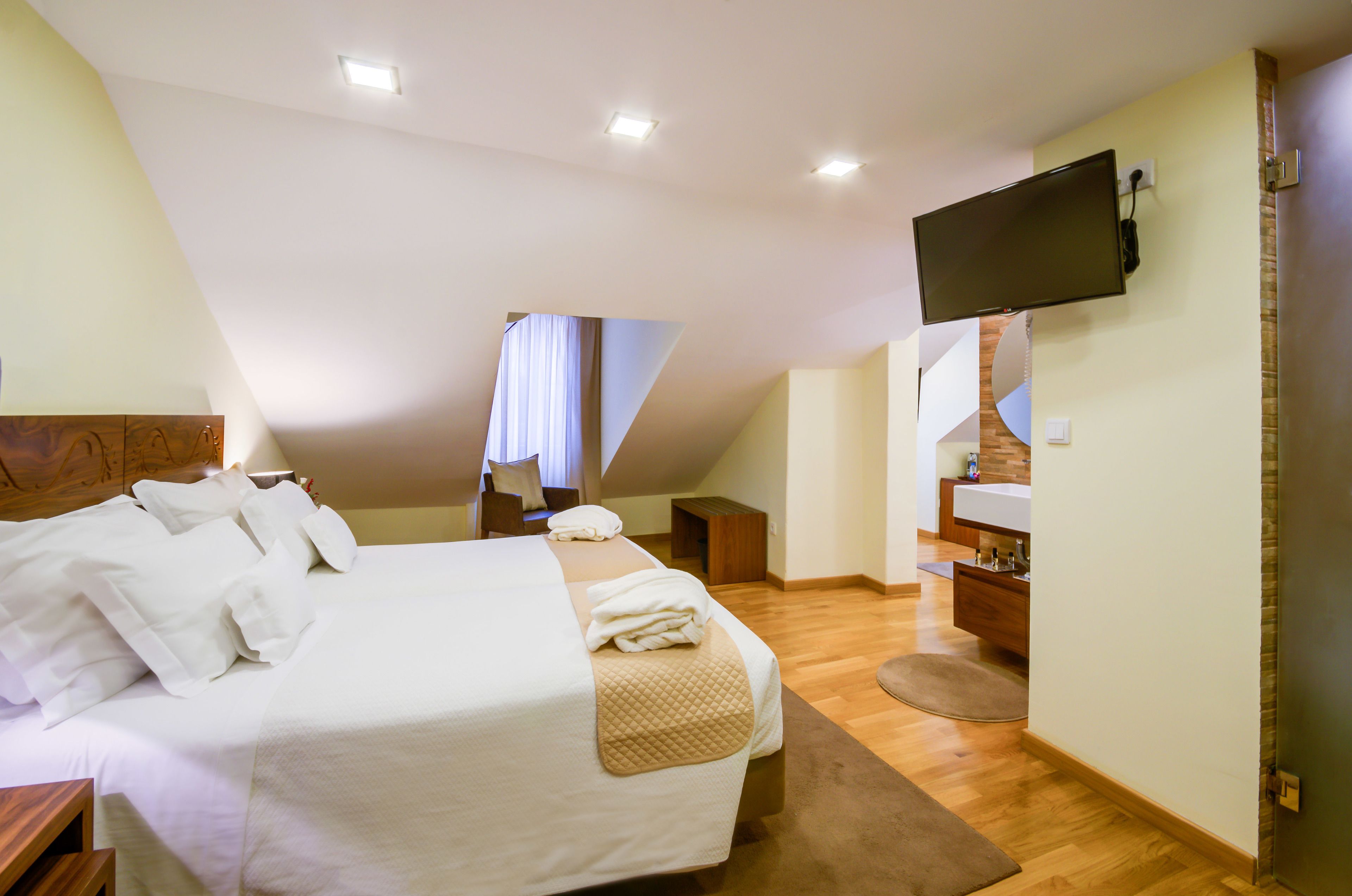 A bedroom with a bed, a TV mounted on the wall, a wooden bench, and a small bathroom area.