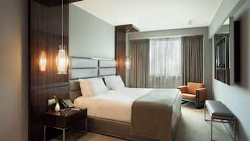 A modern hotel room with a large bed, bedside tables, a chair, a bench, a TV, and a large window with curtains.