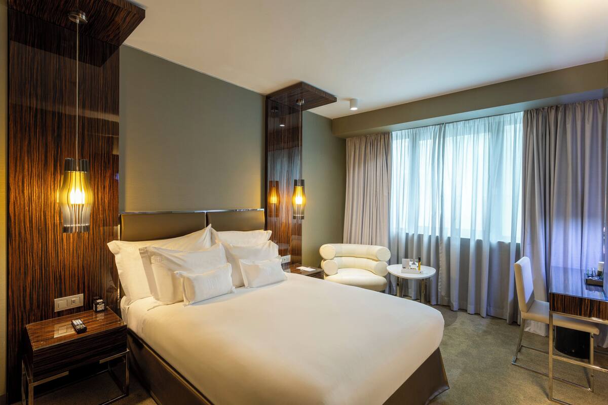 A hotel room with a large bed, a bedside table with a lamp, a chair, a small table, and a window with curtains.