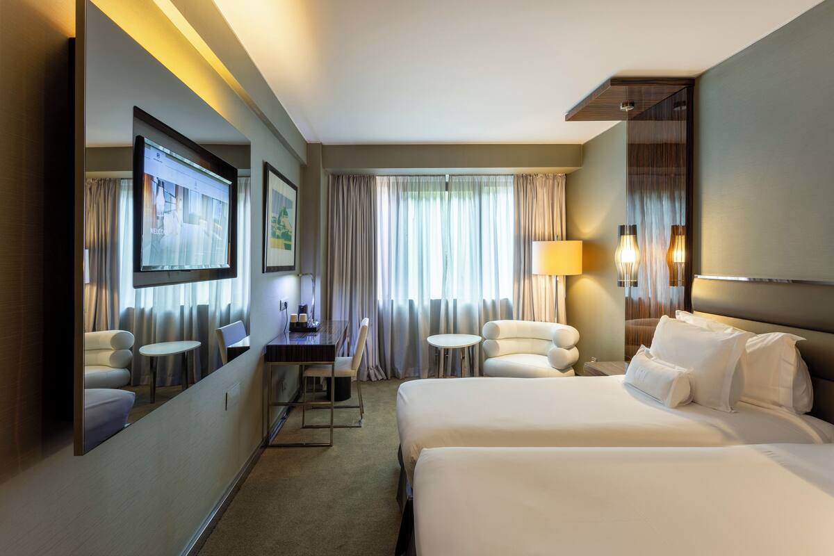A hotel room with two beds, a desk, a chair, a small table, a lamp, and a TV.