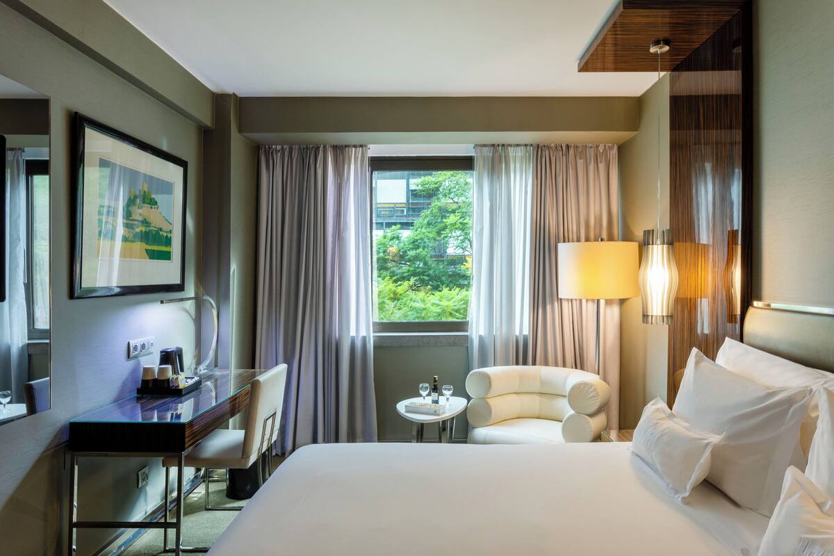 A hotel room with a bed, a desk, a chair, a small table with a bottle and glasses, and a view of greenery outside.