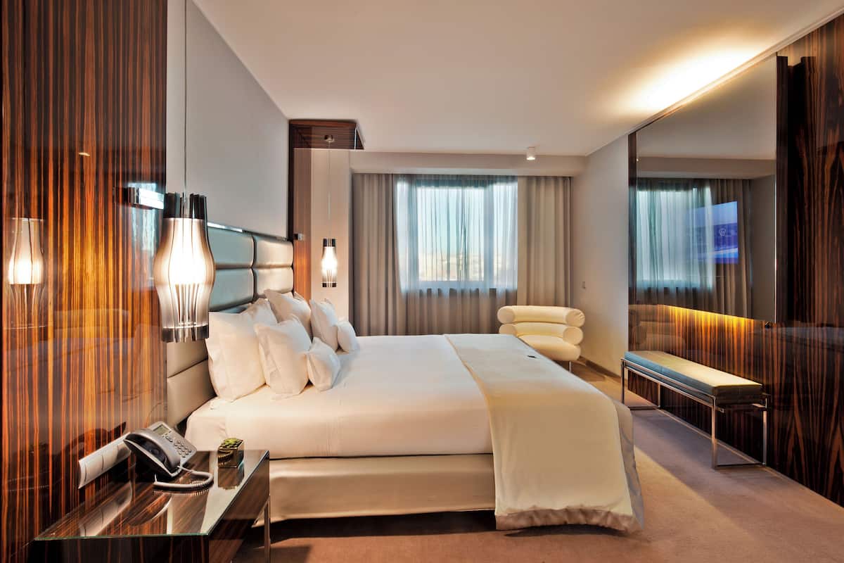 A modern hotel room with a large bed, bedside lamps, a bedside phone, a small table with a lamp, and a bench by the window.