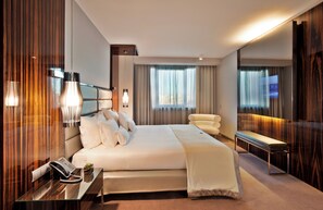 Una habitación de hotel moderna con una cama grande, lámparas de noche, teléfono en la mesita de noche, una pequeña mesa con lámpara y un banco junto a la ventana.
