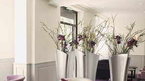 A lobby with a seating area featuring a round table, a white armchair, and purple chairs. There are tall vases with purple flowers and a patterned carpet. - Hôtel Esplanade Eden (Lourdes)