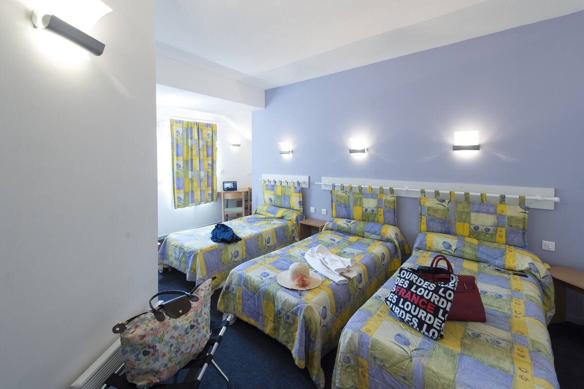 A hotel room with three beds, each with a headboard and a quilted bedspread. There is a window with curtains, a desk with a chair, and a radiator.