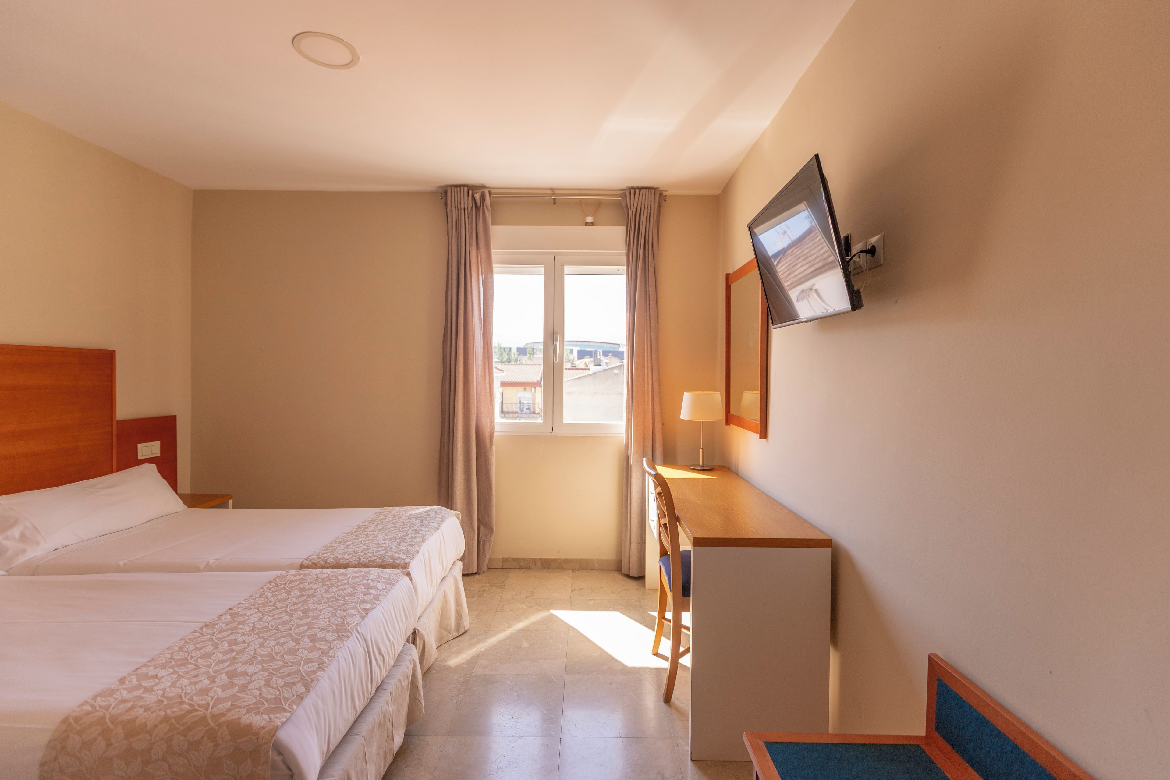 A hotel room with two beds, a desk, a television, and a window with curtains.
