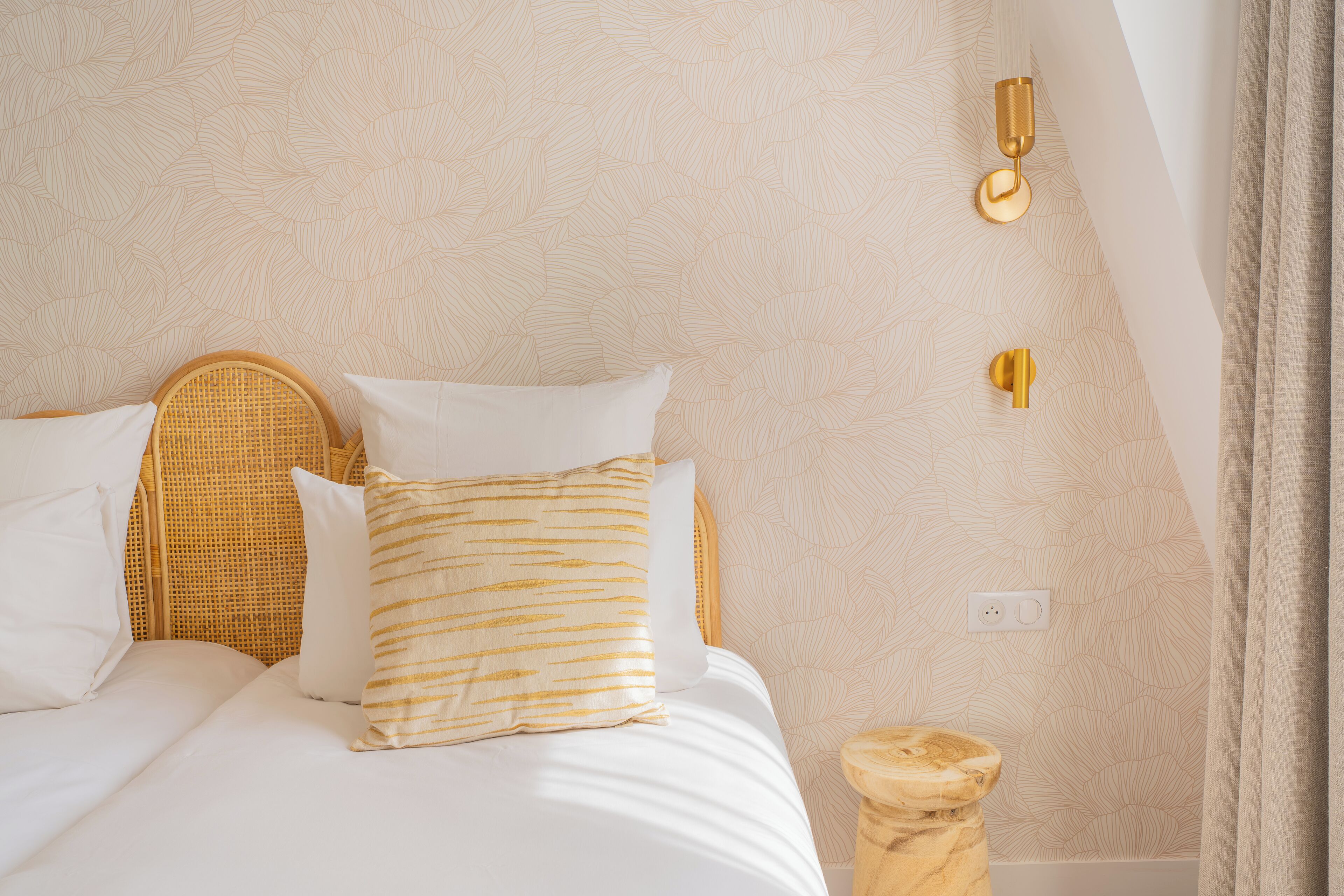 A neatly made bed with white and gold bedding, a wicker headboard, and a small wooden stool beside it.