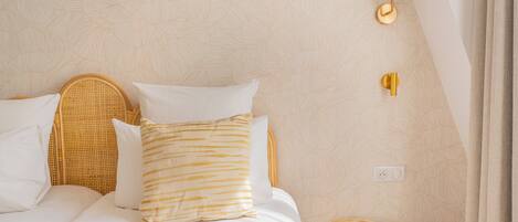 A neatly made bed with white and gold bedding, a wicker headboard, and a small wooden stool beside it.