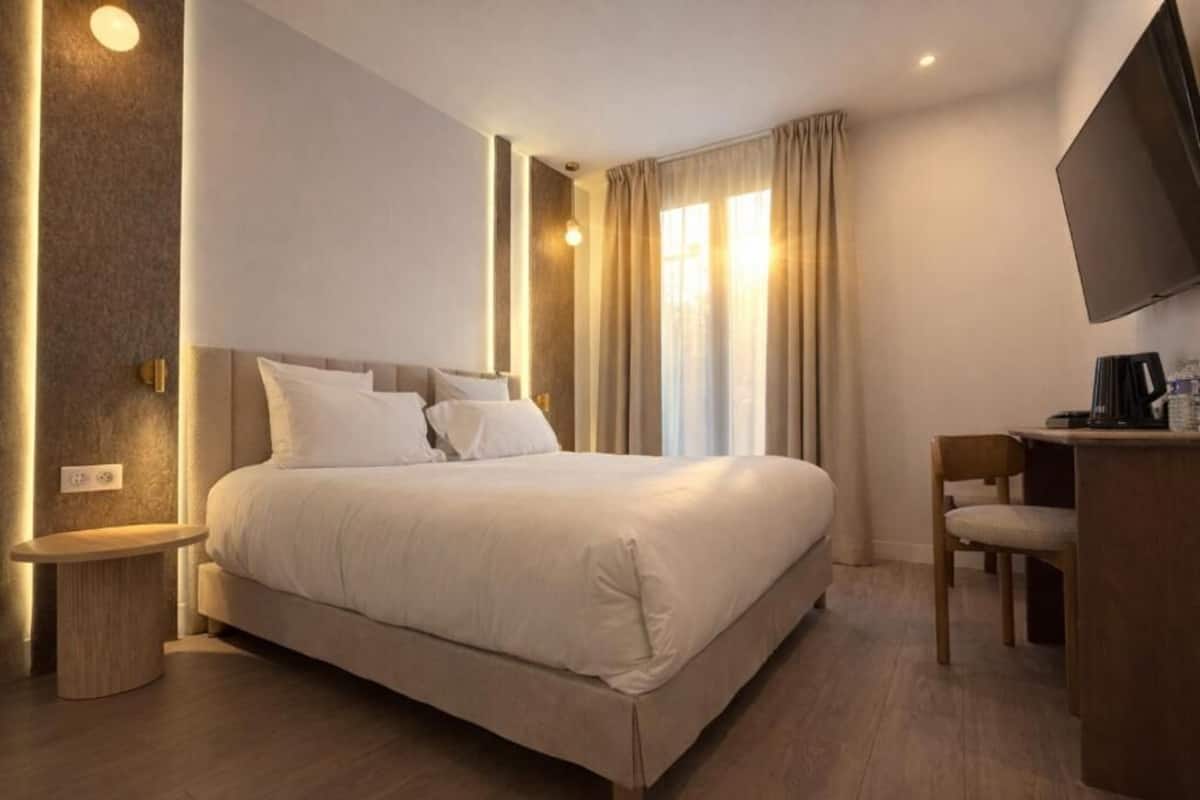 Une chambre d’hôtel comprenant un lit, une table de chevet, une chaise, un bureau, une télévision et des rideaux.