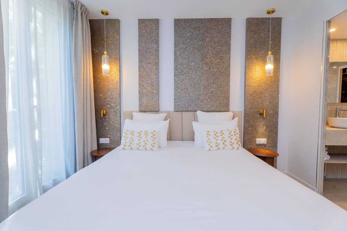 Une chambre d’hôtel moderne avec un grand lit, deux tables de chevet et une salle de bain visible à travers une porte ouverte.