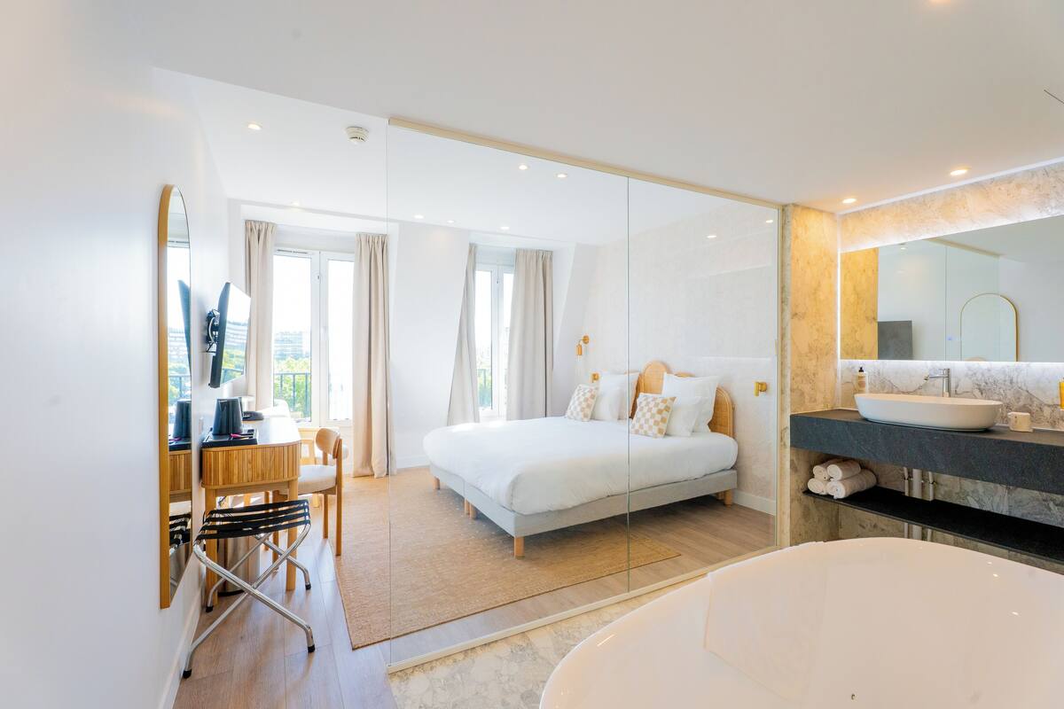 Une chambre d’hôtel moderne dotée d’un grand lit, d’une salle de bain séparée avec une grande baignoire, d’un bureau avec une chaise et d’un grand miroir.