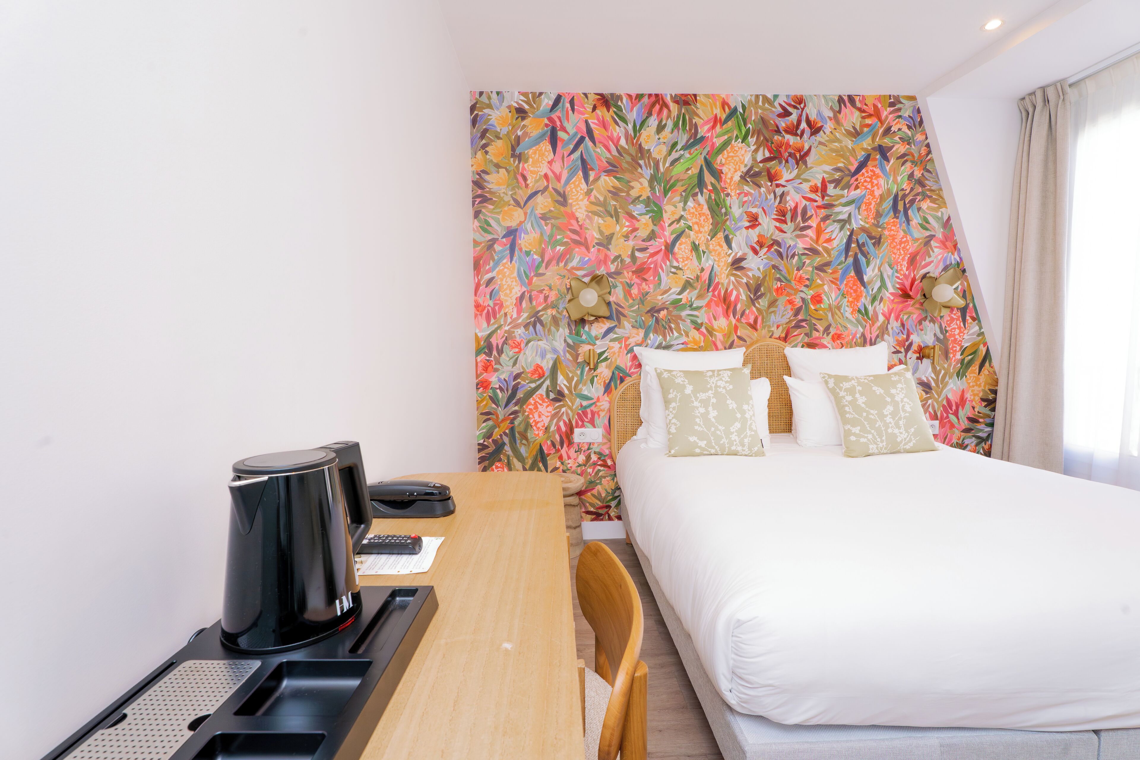 A hotel room with a bed, a desk with a coffee maker, and a colorful floral wall.