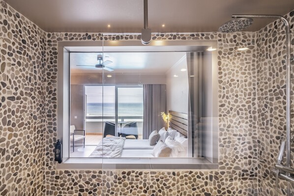 A modern bathroom with a stone wall design, a glass shower enclosure, and a view of a bedroom with a bed and a table. - Arribas Sintra Hotel (Sintra)
