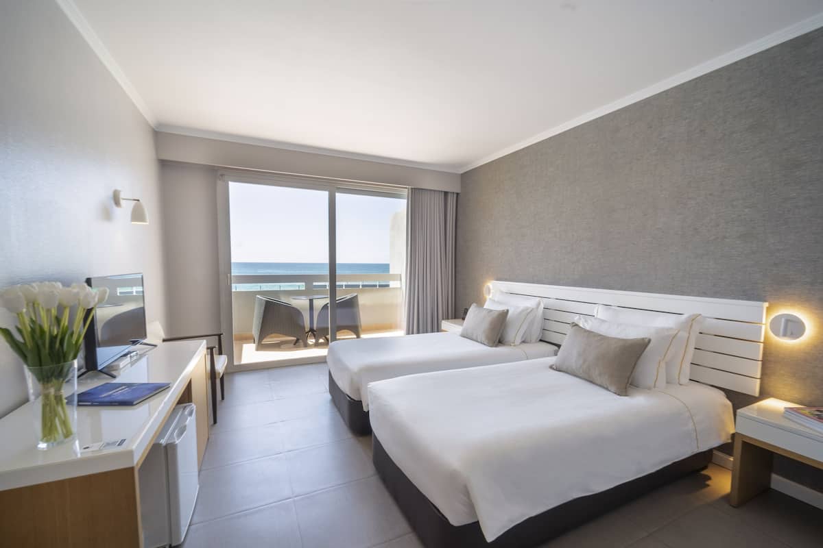 Een hotelkamer met twee bedden, een bureau met een computer, een stoel en uitzicht op zee.