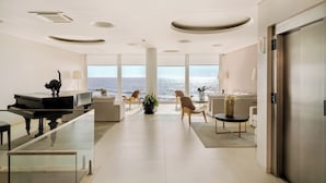 Een moderne hotel lobby met een piano, zithoek, grote ramen met uitzicht op zee, en een minimalistisch ontwerp.