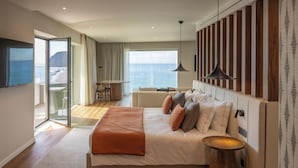 A modern hotel room with a large bed, a balcony with a view of the sea, and a dining area with a table and chairs.