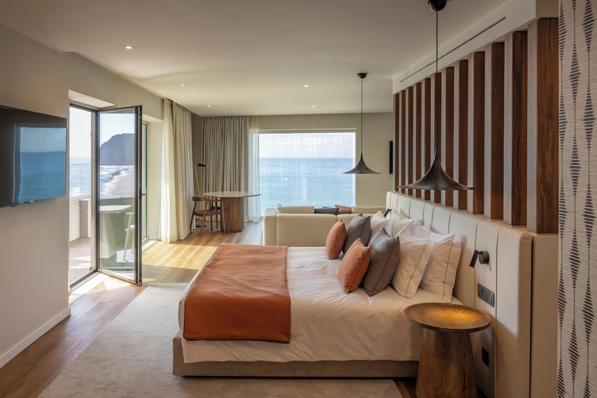 Een moderne hotelkamer met een groot bed, een balkon met uitzicht op zee en een zithoek met een tafel en stoelen.
