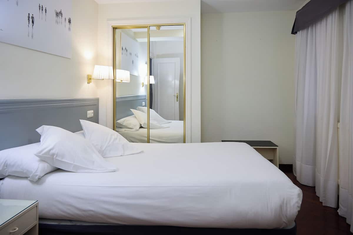 A hotel room with a bed, a nightstand, a lamp, a mirror, and a door with a view of the hallway.