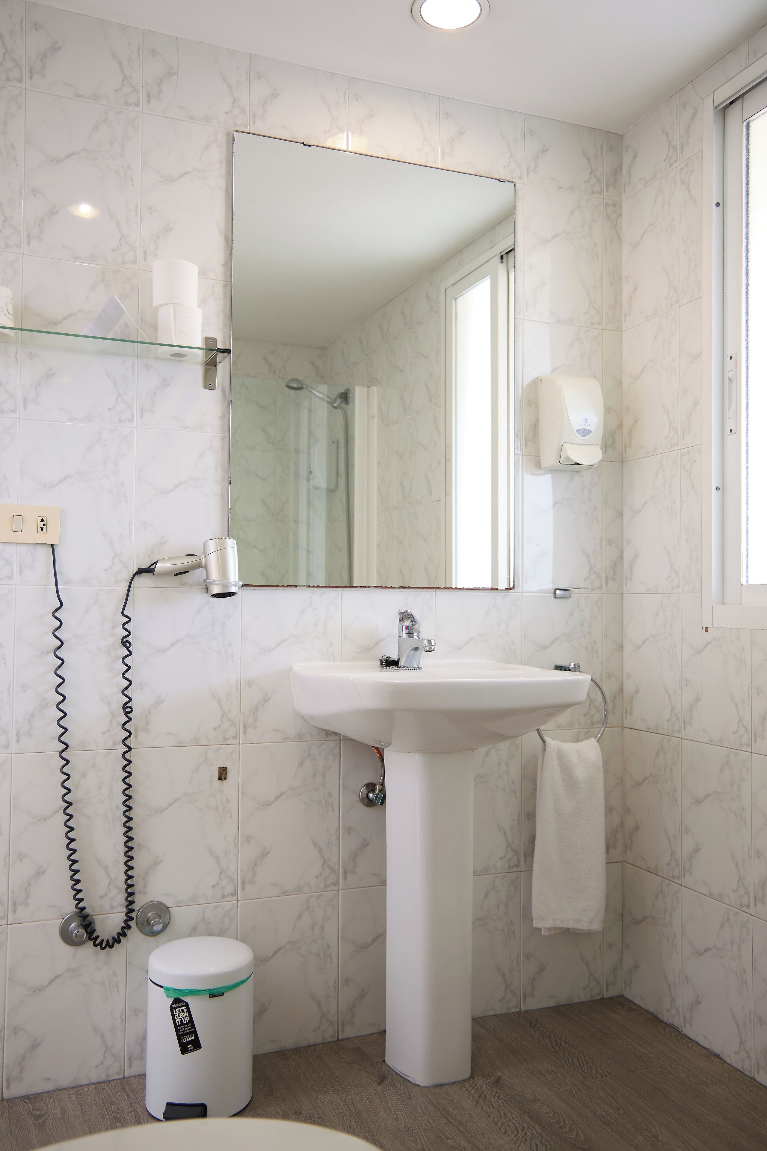 Een badkamer met een witte wastafel, een spiegel, een handdoek aan de muur en een afvalbak.