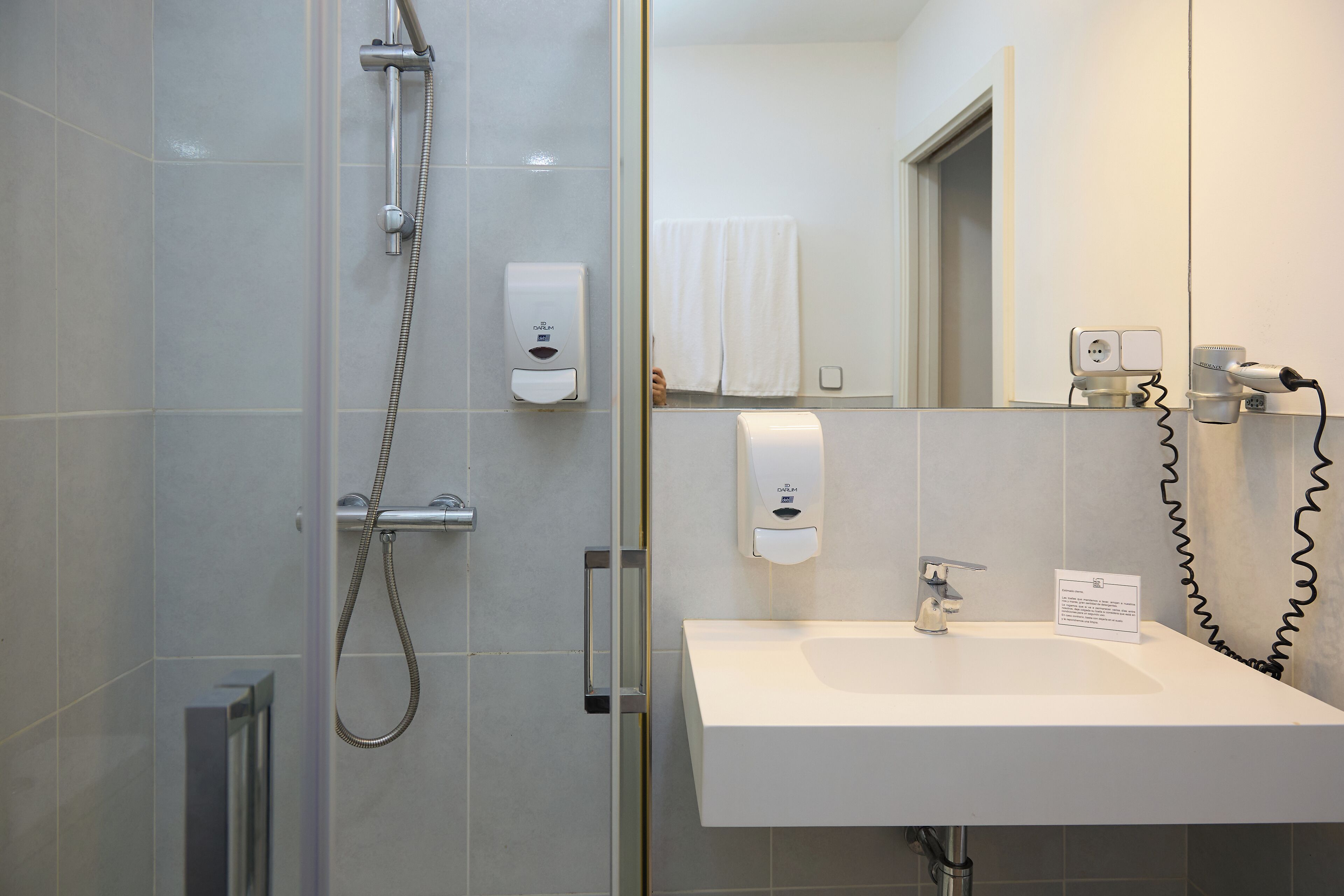 Un baño moderno con cabina de ducha de vidrio, lavamanos blanco y dispensador de toallas.