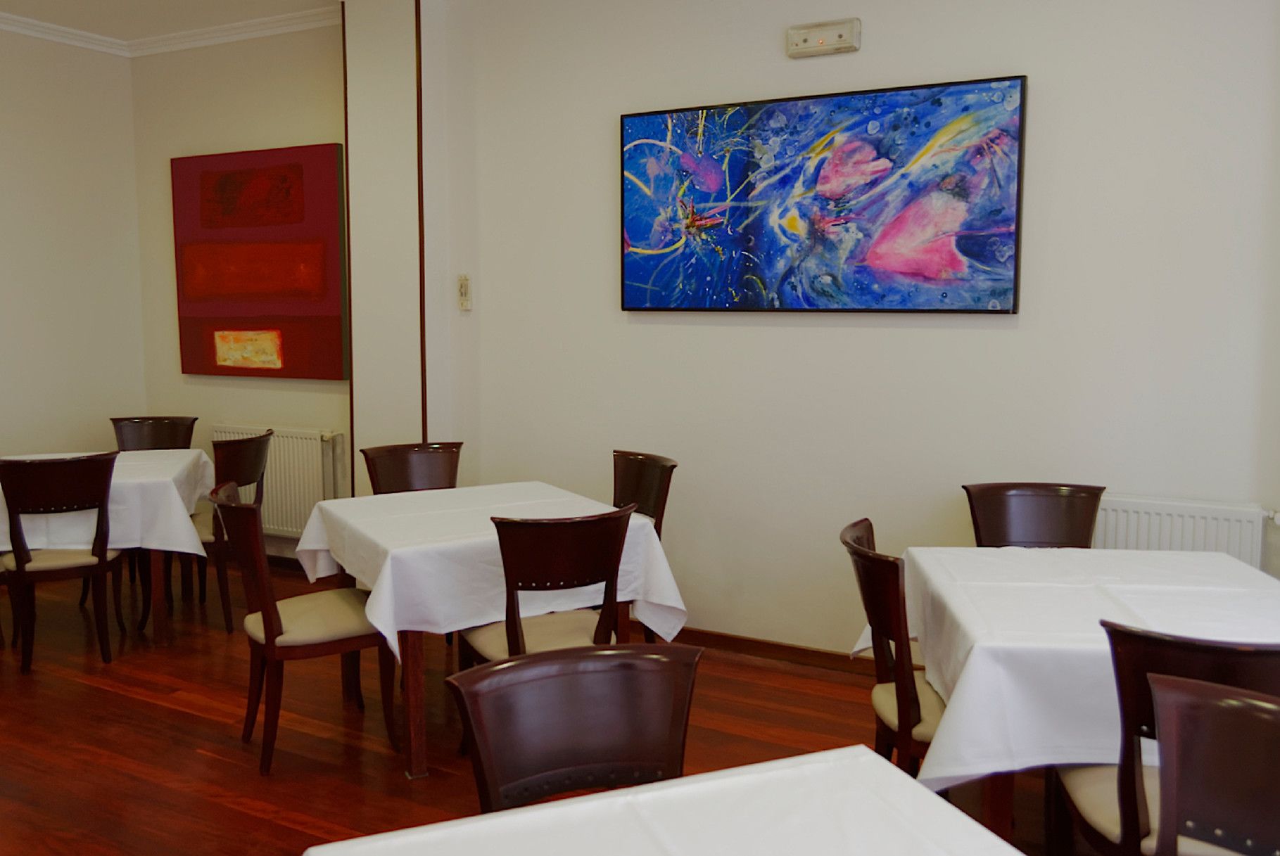 A dining area with tables and chairs, a large abstract painting on the wall, and a red artwork below it.
