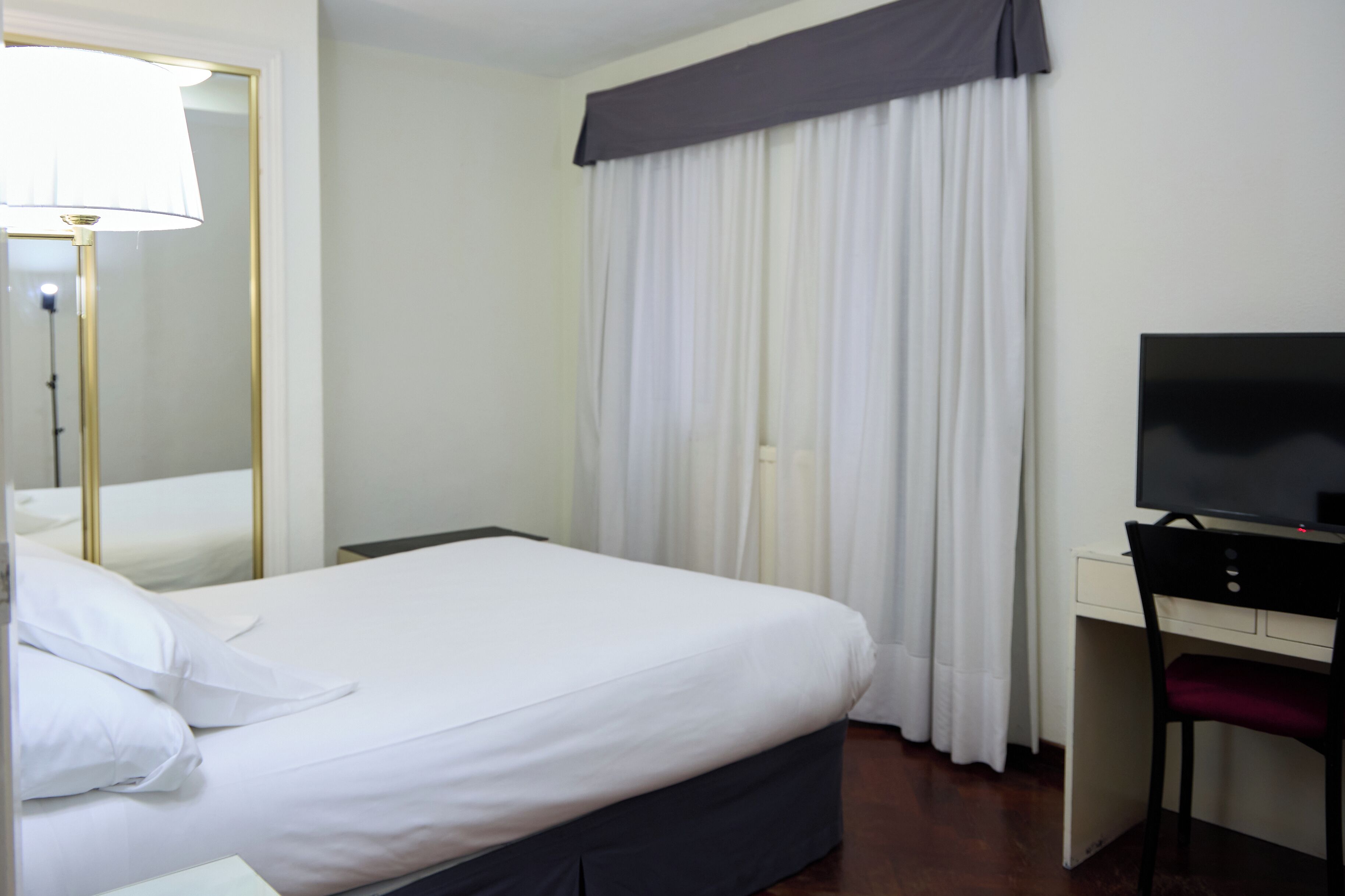 A hotel room with a bed, a desk, a chair, a television, and a window with curtains.