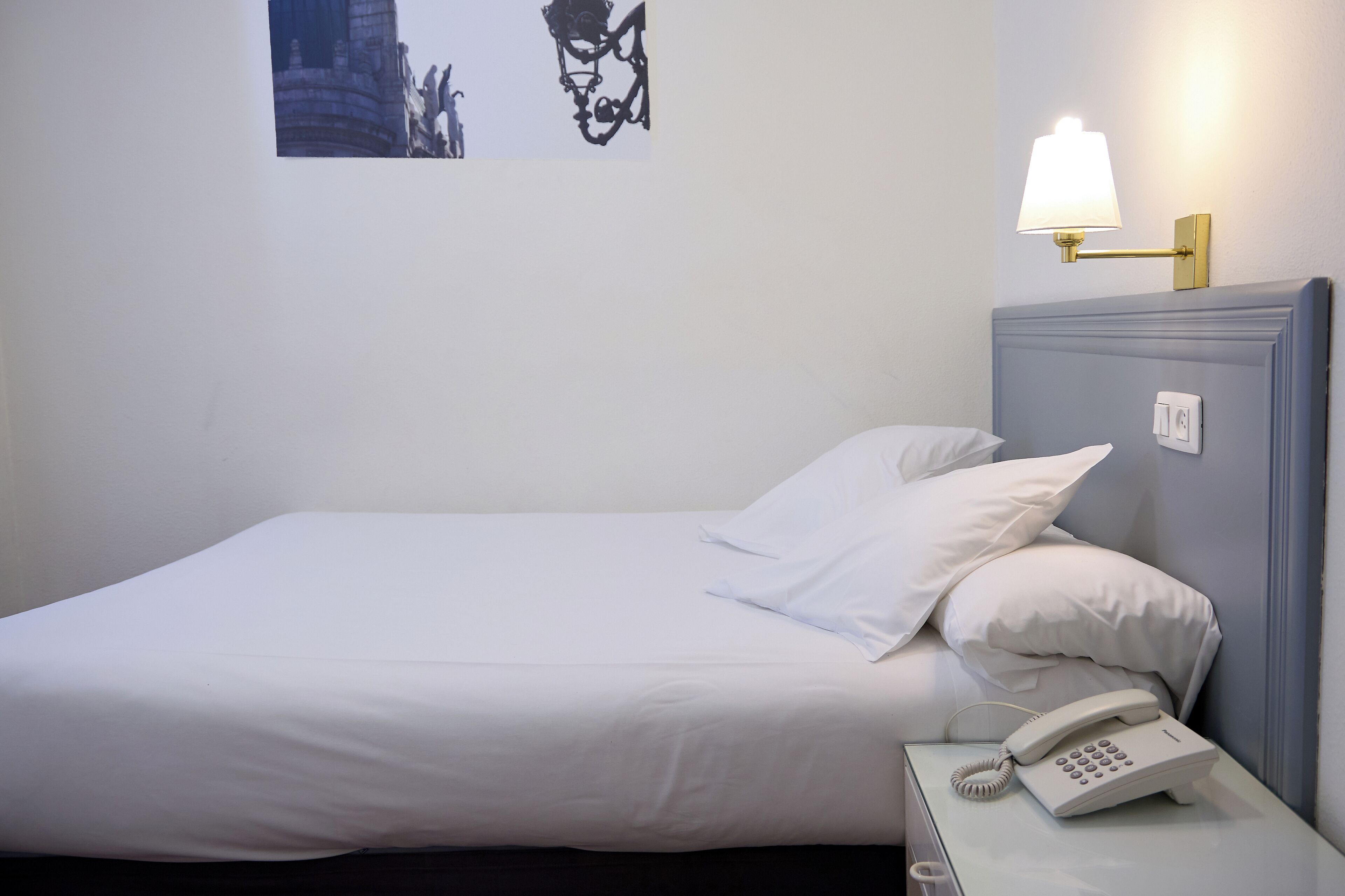 Una cama bien hecha con sábanas blancas, una lámpara de noche y un teléfono en la mesita de noche.