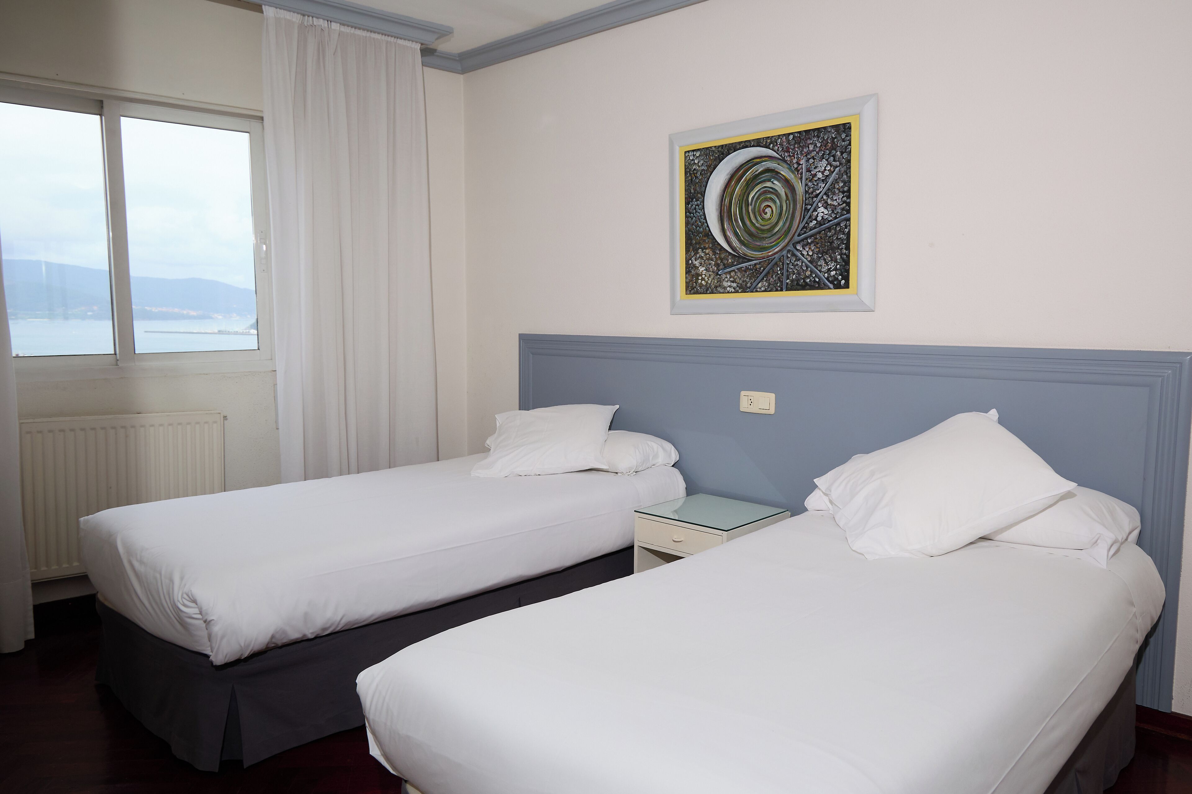 Habitación de hotel con dos camas individuales, una mesita de noche, una ventana con cortinas y un cuadro abstracto enmarcado en la pared.