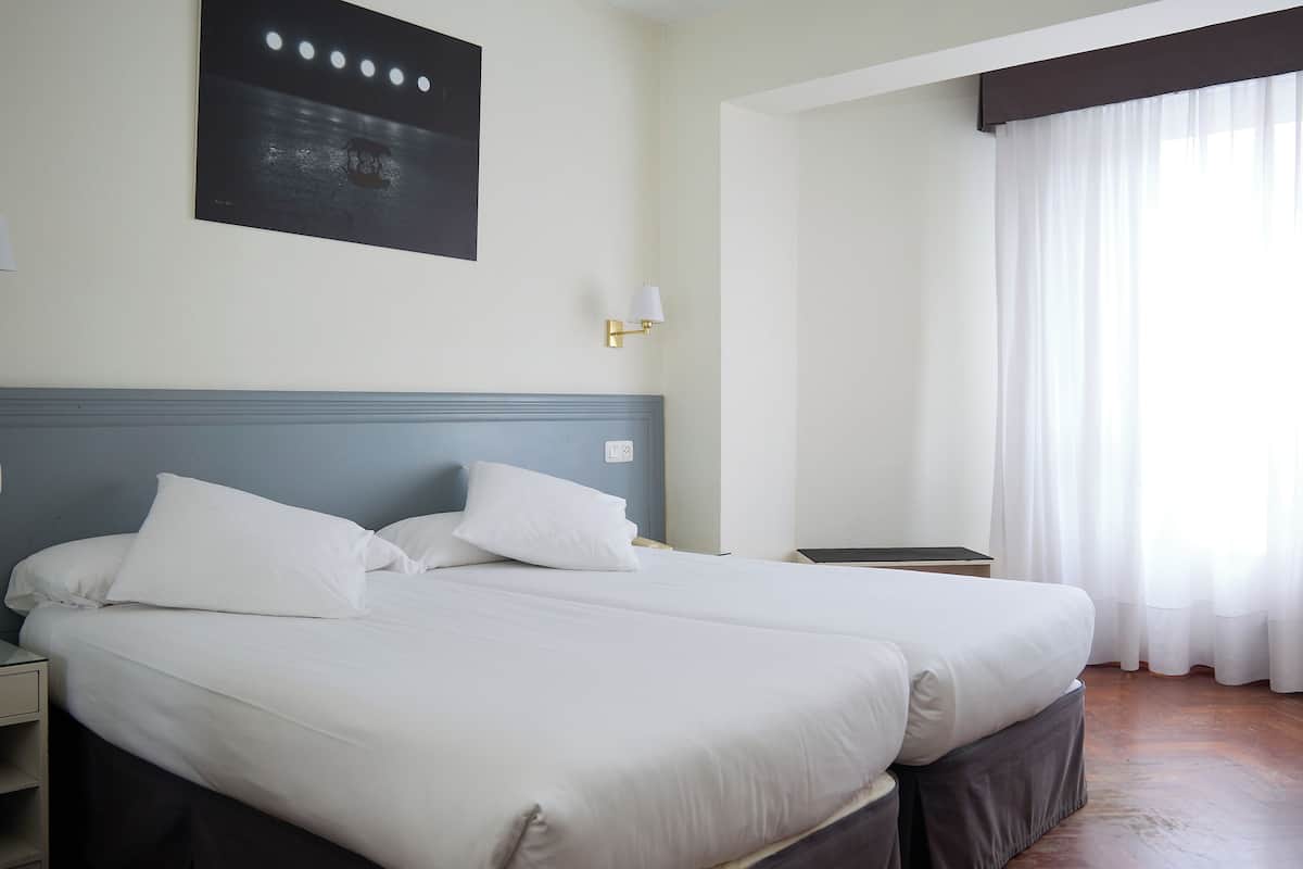 A hotel room with two beds, a nightstand, a wall-mounted lamp, and a window with curtains.