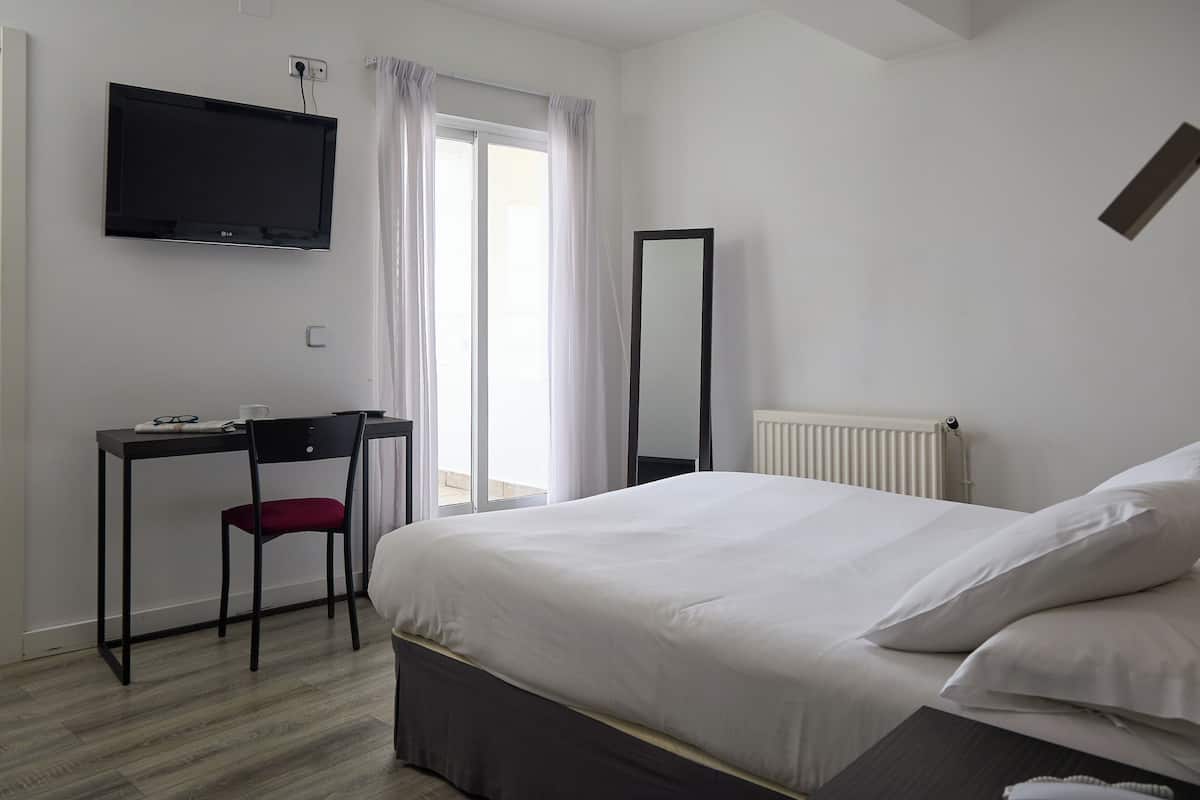 A hotel room with a bed, a desk with a chair, a television mounted on the wall, and a window with curtains.