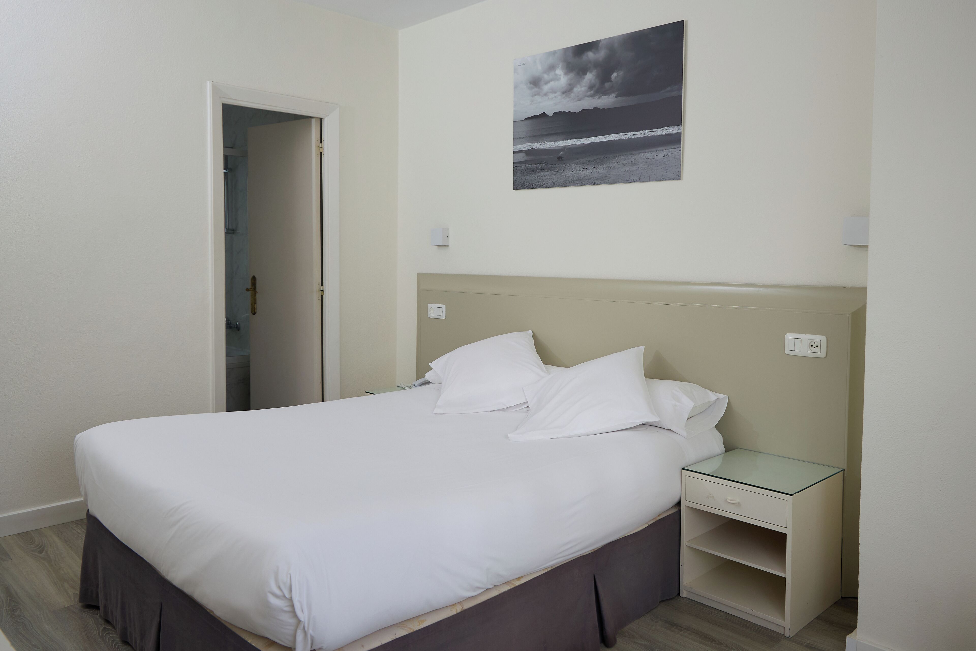Un lit bien fait, avec du linge de lit blanc, dans une pièce dont le mur est de couleur beige et où est accrochée une image encadrée représentant une scène de plage.