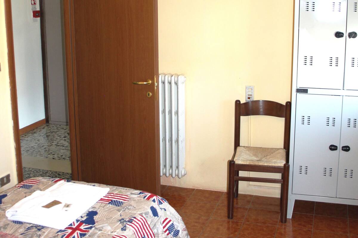 Une chambre d’hôtel comprenant un lit, une chaise en bois, un radiateur, un téléviseur et un ensemble de casiers.