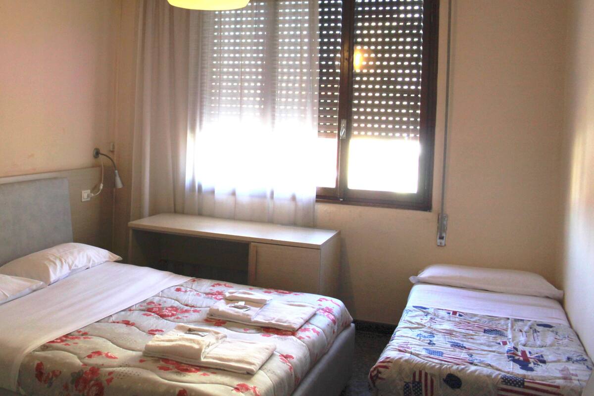 Une chambre d’hôtel avec un lit simple, un petit bureau et une fenêtre avec des stores.